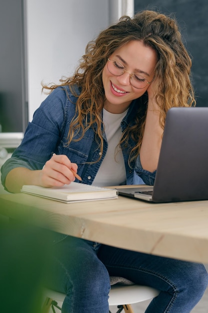 Young female in casual clothes sitting at table with laptop and working on project while taking notes in planner