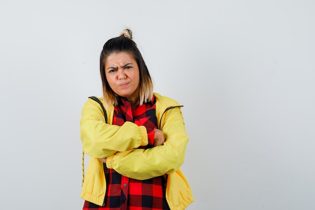 Young female in checkered shirt, jacket holding arms folded, curving lips and looking disappointed , front view.