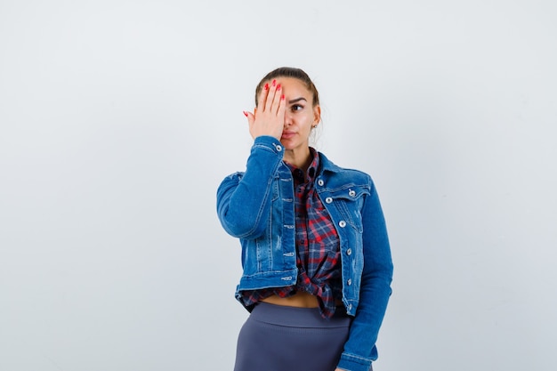 Young female in checkered shirt, jacket, pants with hand on eye and looking serious , front view.