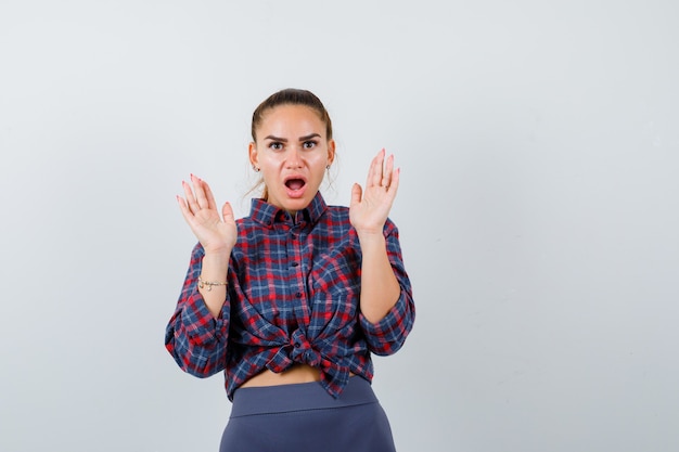 Young female in checkered shirt, pants showing palms in surrender gesture and looking puzzled , front view.