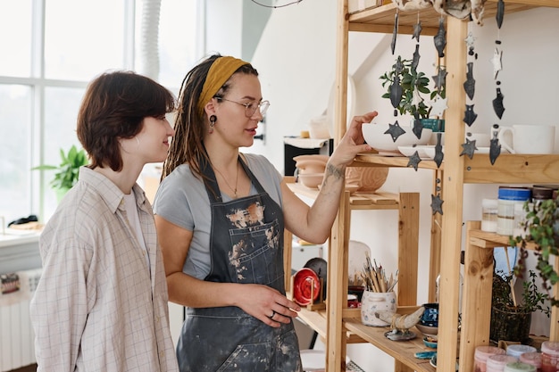 Young female clerk of small earthenware shop showing new items to customer