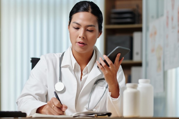 Young female clinician in lab coat making notes during phone call