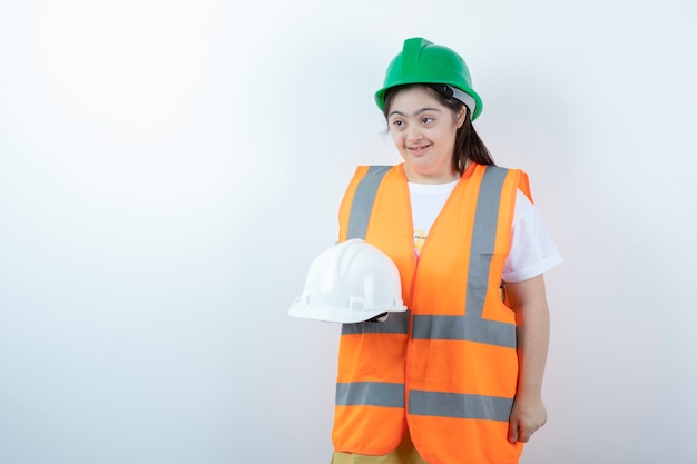 Young female construction worker in hardhat holding helmets over white wall. 