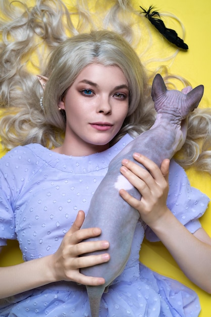 Young female cosplay elf in blue dress looking at camera lying down with cat on yellow background