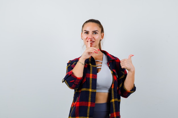 Young female in crop top, checkered shirt, pants showing silence gesture while pointing thumb up to the right side and looking pretty , front view.