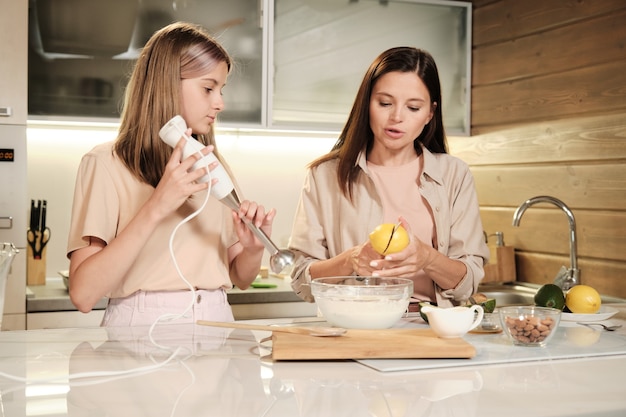 Young female cutting lemon into two halves over bowl with milk while her daughter is going to mix the ingredients by electric mixer