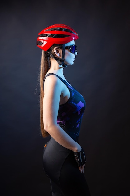 A young female cyclist wearing a safety helmet and glasses dressed in a bib shorts poses against a black background in the studio