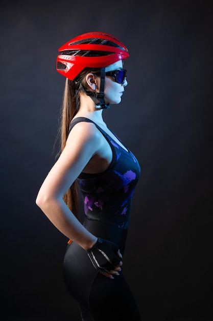 A young female cyclist wearing a safety helmet and glasses dressed in a bib shorts poses against a black background in the studio