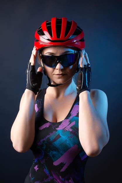 A young female cyclist wearing a safety helmet and glasses dressed in a bib shorts poses against a black background in the studio