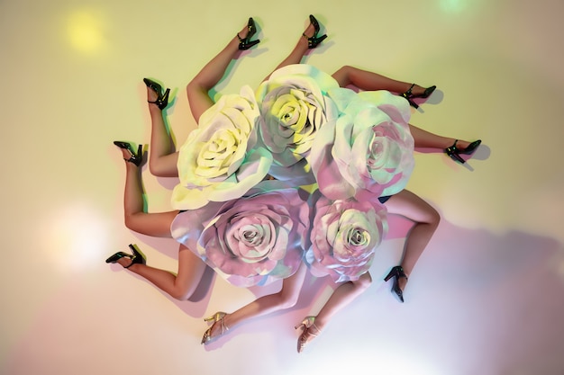 Young female dancers with huge floral hats in neon light on gradient wall