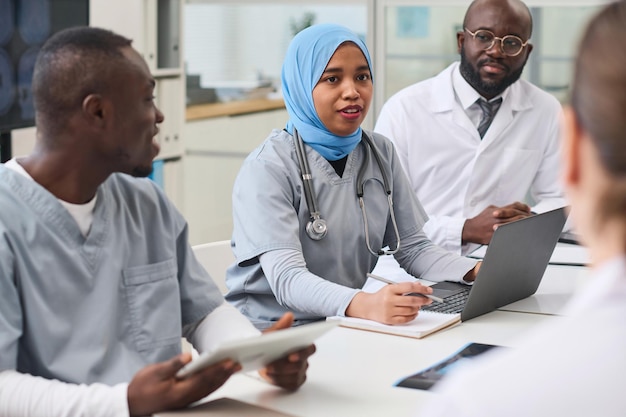 Young female doctor in hijab using laptop and talking to her colleagues during conference in office