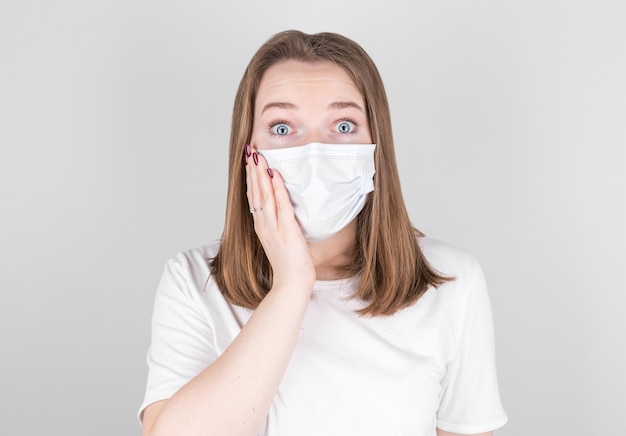 Young female doctor in a medical mask looks into the camera with surprise, holding her face with her hand. after contracting COVID 19, flu and seasonal colds.