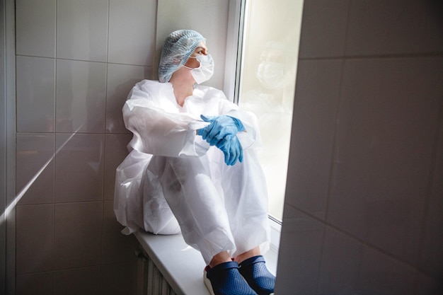 A young female doctor in a protective suit cap mask and gloves is sitting wearily near the window on...