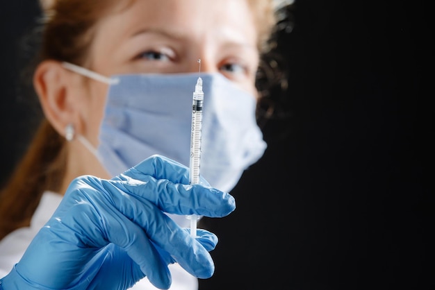 A young female doctor with a syringe looks at the camera a nurse wearing a medical mask and blue lat