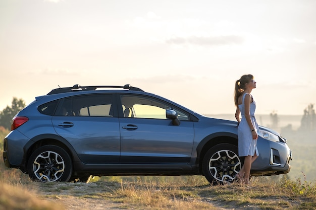 Young female driver resting near her car enjoying warm summer evening. Travel and getaway concept.