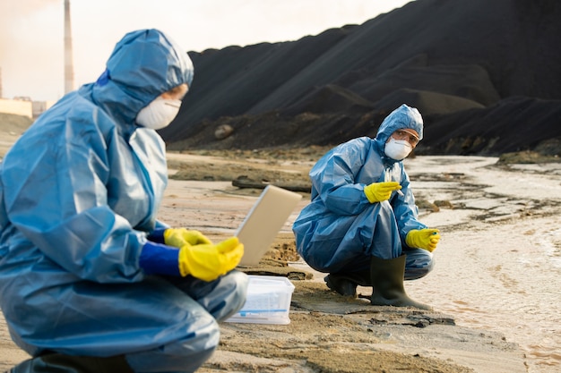 Young female ecologist holding sample of dirty water while looking at her colleague entering data about its characteristics in dangerous area