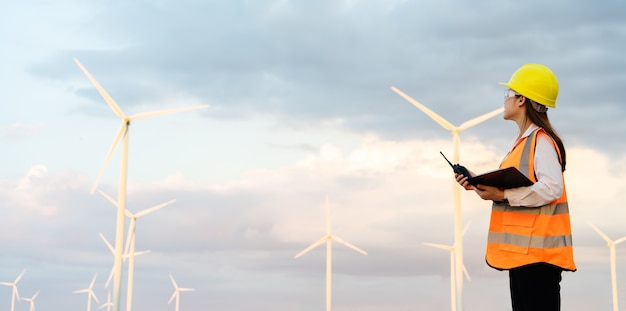 Young female engineer using walkie talkie and reading clipboard to checking system against wind turbine farm