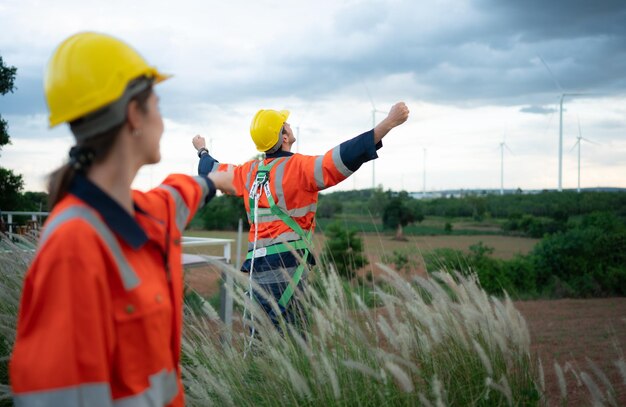 Photo young female engineer working in a wind turbine field and young man engineer wear a safety vest raise both hands to relax after finishing the wind turbine inspection mission