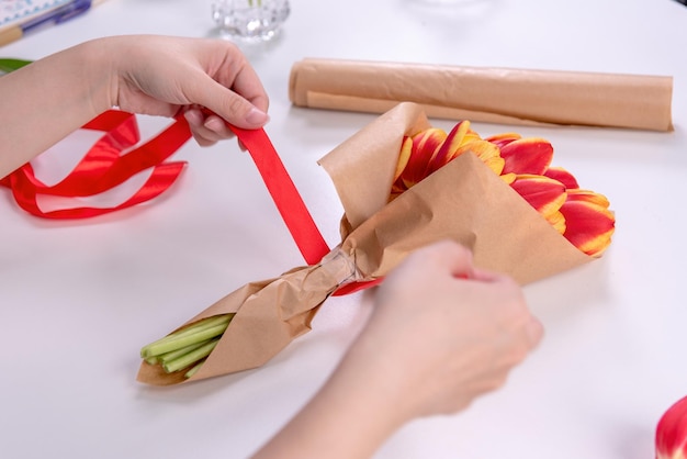 Young female florist is wrapping a red color tulip flower bouquet for Mother's Day gift with tie ribbon bow over a white table background close up lifestyle