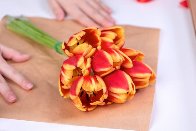 Young female florist is wrapping a red color tulip flower bouquet for Mother's Day gift with tie ribbon bow over a white table background, close up, lifestyle.