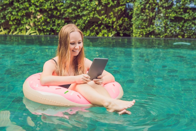 Young female freelancer sitting near the pool with her laptop in the hotel browsing in her smartphone. Busy at holidays. Distant work concept. Copy space for your text