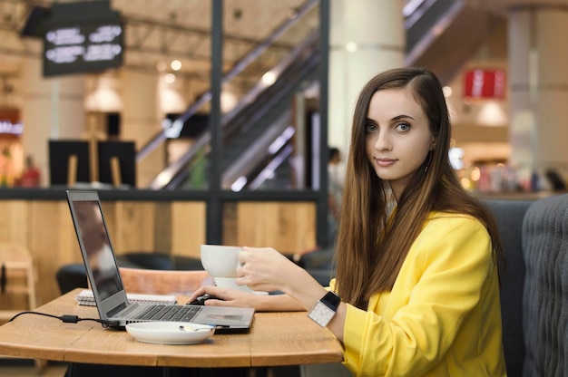 Young female freelancer in yellow jacket working in a cafe with laptop and drinking coffee