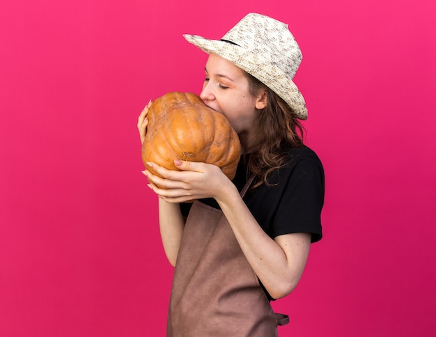 Young female gardener wearing gardening hat holding bites of pumpkin isolated on pink wall