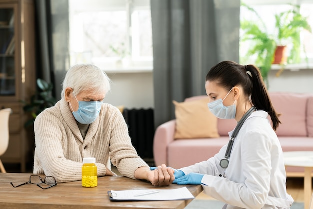 Young female general practitioner in whitecoat and protective mask touching pulse of sick senior male patient during medical check-up