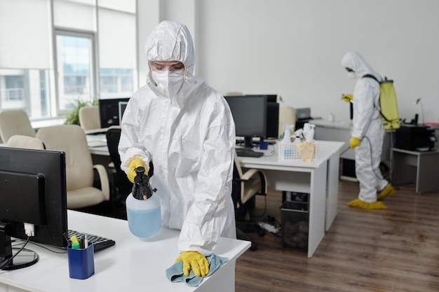 Young female in hazmat suit wiping desks while spraying disinfectant on surface