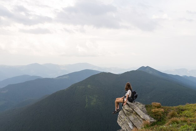 Young female hiker sits on rock in gorgeous mountain area and looks in endless horizon