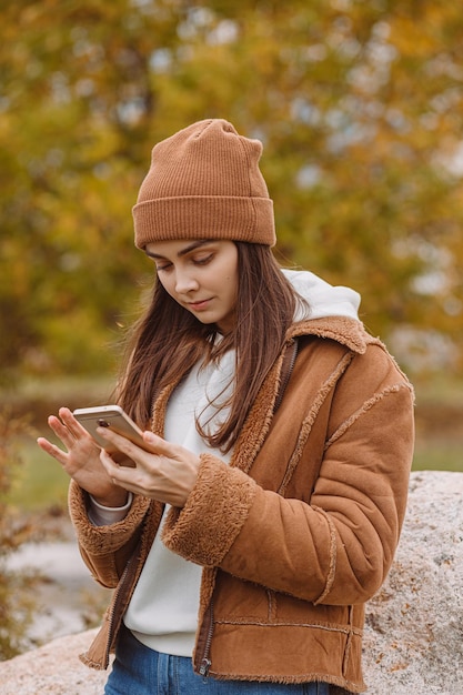 Young female hipster in outerwear using cellular and browsing social media in autumn park