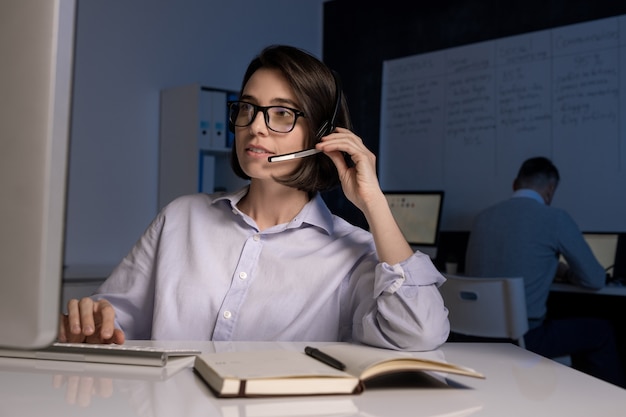 Young female hotline operator with headset speaking to clients online in front of computer screen