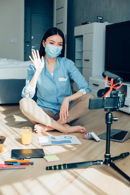 Young female interior designer sitting on the floor in front of a camera and waving her hand Medical mask on her face