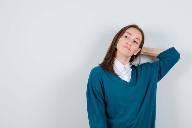 Young female keeping hand behind head, looking away in sweater over shirt and looking focused. front view.