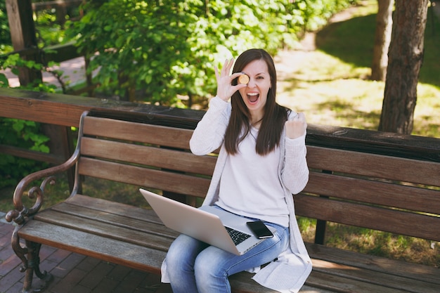 Young female in light casual clothes. Woman sitting on bench holding bitcoin coin of golden color working on modern laptop pc computer in street outdoors. Mobile Office Online virtual currency concept