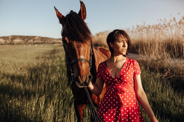 Young female in long red dress with horse looking away while standing on yard of ranch on sunny day in countryside