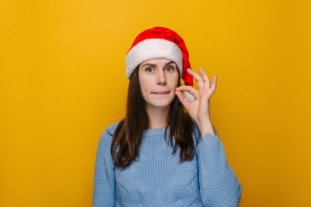 Young female losing her mouth with zip gesture as promise to keep secret rumor, dressed in Christmas hat and winter sweater, isolated on pastel yellow studio background. Happy New Year concept