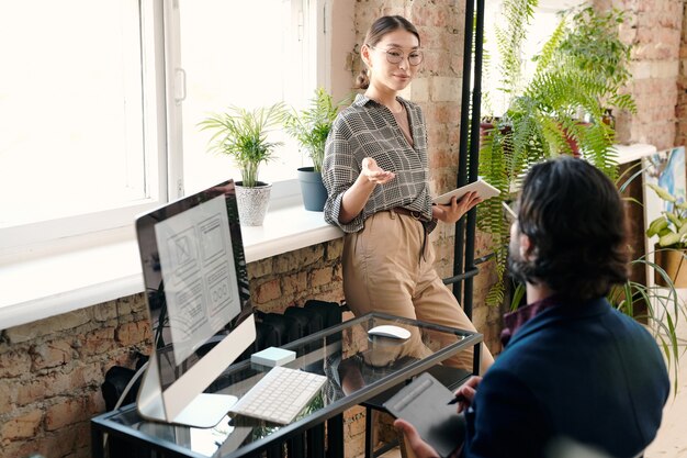 Young female mobile application developer with tablet having discussion with her male colleague drawing sketch in front of computer screen