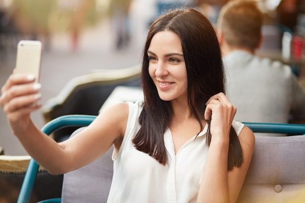 Young female model with dark straight hair and gentle smile poses for making selfie, sits against outdoor sidewalk cafe, uses modern smart phone. Positive delighted European woman in restaurant