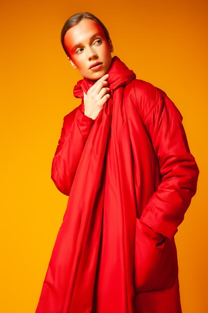 Young female modern wrapping in warm oversize red coat and looking at camera against yellow background