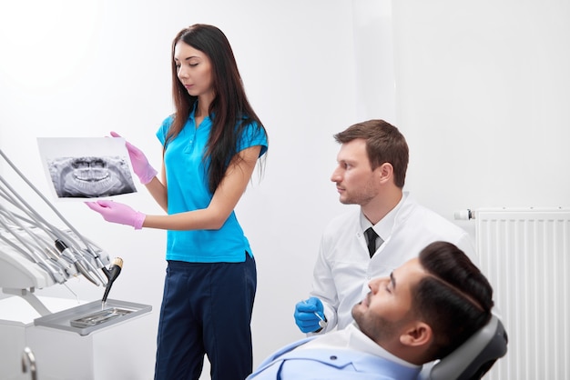 Young female nurse holding x-ray scan of a jaw assisting mature male