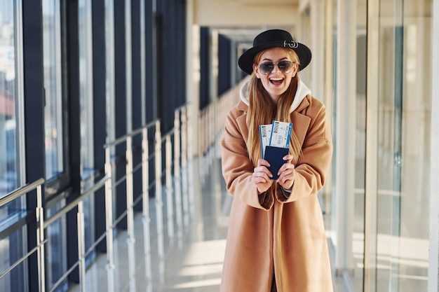 Young female passenger in warm clothes showing tickets in airport hall.