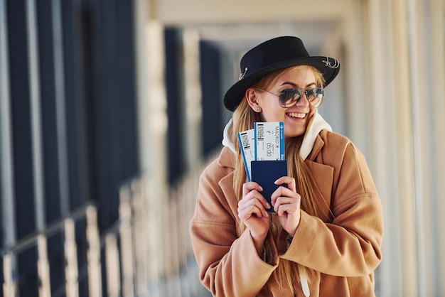 Young female passenger in warm clothes showing tickets in airport hall.