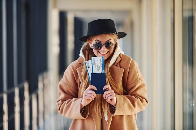 Young female passenger in warm clothes showing tickets in airport hall.