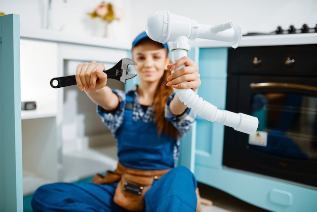 Young female plumber in uniform shows wrench and pipe in the kitchen. Handywoman with toolbag repair sink, sanitary equipment service at home