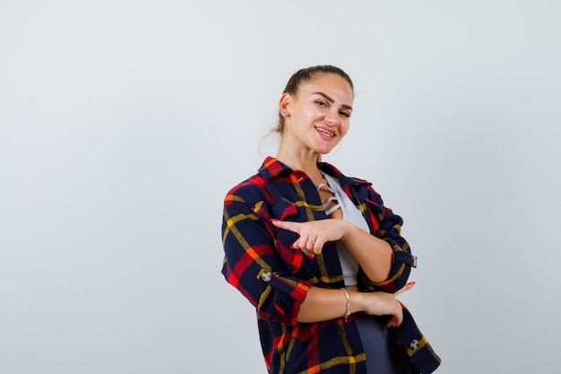 Young female pointing to the left side in crop top, checkered shirt, pants and looking happy. front view.