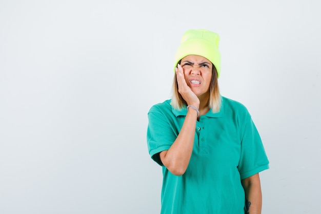 Young female in polo t-shirt, beanie suffering from toothache and looking painful , front view.