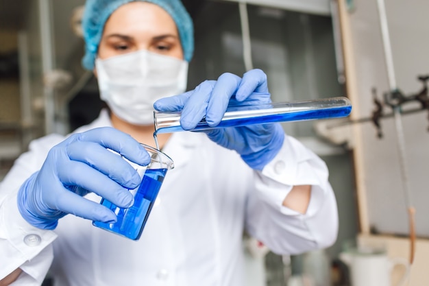 Young female researcher in medical cap and protective mask are doing investigations with test tube