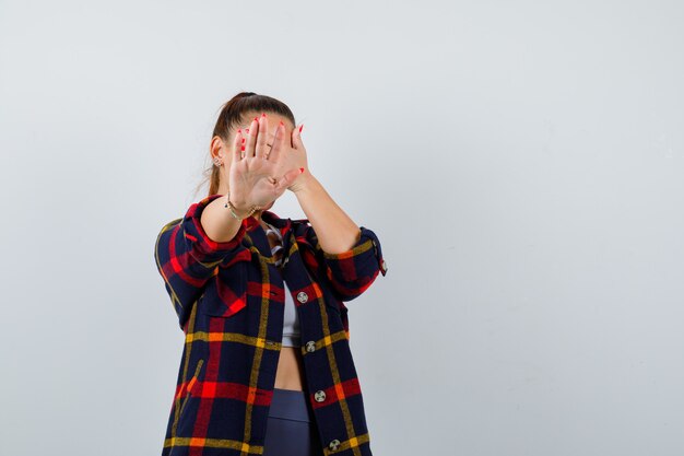 Young female showing stop gesture, covering eyes with hand in crop top, checkered shirt and looking serious. front view.