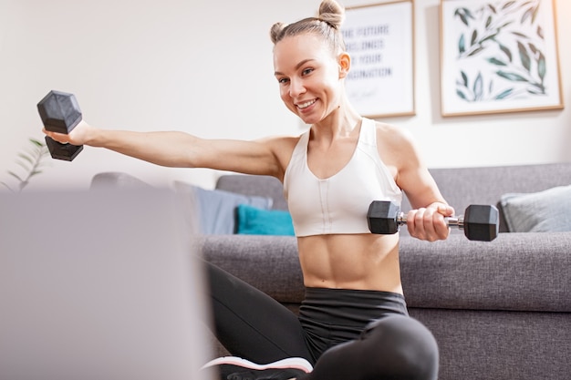 Young female smiling and lifting dumbbells while watching fitness course on laptop during self isolation at home